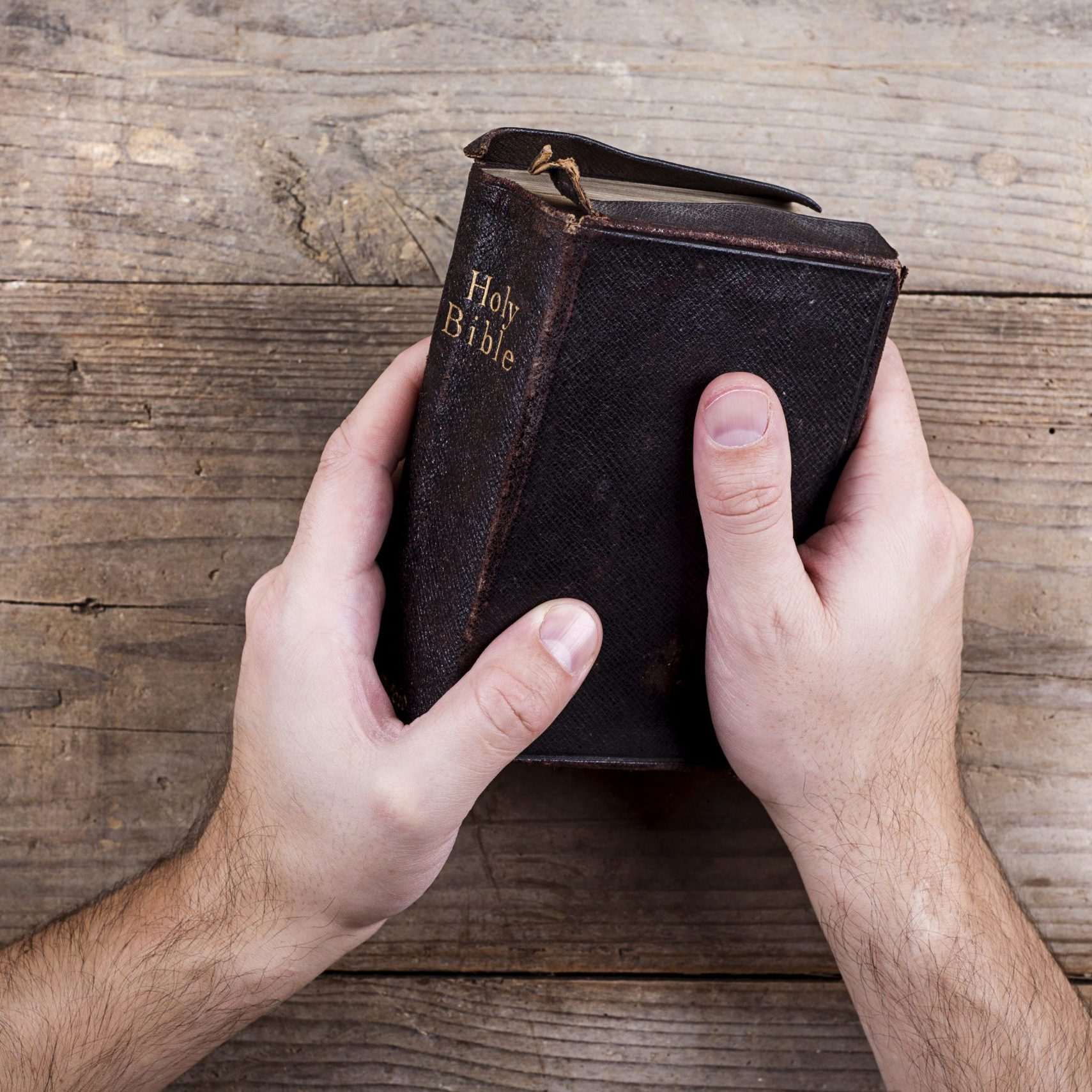 Hands holding Bible on a wooden desk background.